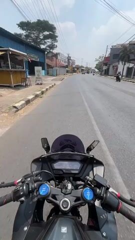 The video shows a first-person perspective of someone riding a motorcycle on a paved road. The motorcycle is black with red accents. The rider's hands are visible on the handlebars, and the digital dashboard of the motorcycle is in the foreground. The road appears to be in a somewhat urban or suburban area, with buildings and trees lining the sides. There are other vehicles and structures visible in the distance. The sky is bright and appears to be daytime. The video progresses with slight camera movements as the motorcycle moves forward.