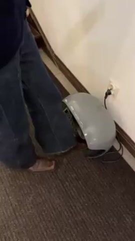 A person wearing jeans and brown dress shoes is operating a gray-colored foot massager. The massager is plugged into a wall outlet and positioned on a striped brown and beige rug next to a white wall. The person is seen placing their feet inside the foot massager, which is running. The setting appears to be in a hallway near a staircase with decorative tiles.
