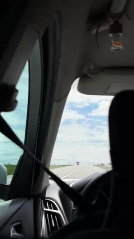 The view from the driver's seat of a car shows a highway with a truck ahead. The sky is visible through the windshield, with scattered clouds. The driver is wearing a seatbelt. The interior of the car is visible, including the dashboard, air vents, and rear-view mirror. There is music playing.