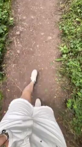 The video shows a first-person perspective of someone walking on a dirt path, surrounded by dense green vegetation. The person is wearing white shorts and white shoes. Another person walks slightly ahead on the path. The path is narrow and winding, and the surrounding foliage is lush and overgrown. The camera pans upward to show the dense canopy of trees overhead, creating a shaded environment. The overall impression is a peaceful walk in a natural setting.