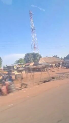 The video shows a view from a moving vehicle of a rural landscape. In the foreground is a paved road. Adjacent to the road, the landscape is a reddish-brown dirt. There are structures, trees, and a tall communication tower in the background. The tower has a red and white painted pattern. Several buildings are visible, and they appear to be simple, possibly informal structures. There are also piles of what seems to be chopped wood in one area. The sky is blue and mostly clear. A blue van passes on the side of the road. There are a few people walking around in the distance, but they are not clearly visible. The overall impression is of a rural or developing area. The camera is shaky due to the movement of the vehicle.