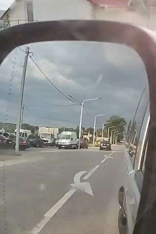 The video shows a view through a car's side mirror of a road with a cloudy sky overhead. On the left side of the road, there is a building with a red roof and a pile of construction debris. Several vehicles are on the road, including a white van and other cars. Streetlights line the right side of the road. The car appears to be moving, as indicated by the slight blur of the surrounding environment.