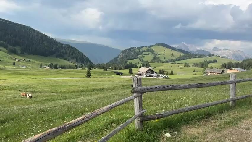 This media file captures a picturesque mountain landscape in Kastelruth - Castelrotto, Italy. The scene is dominated by rolling green hills, lush with grass and dotted with wildflowers. In the foreground, a rustic wooden fence made of rough-hewn logs and posts separates the viewer from the expansive meadow.

Beyond the fence, several buildings are nestled into the landscape. A prominent structure with a white facade and dark roof, resembling a farmhouse or lodge, stands prominently. To its right, a smaller, traditional wooden building with a similar dark roof adds to the pastoral charm. In the distance, more scattered buildings and the dark green of pine forests climb the slopes of the mountains. The background is defined by the majestic peaks of the Dolomites, their rugged outlines softened by the atmospheric conditions.

The activity taking place is seemingly tranquil. Two cows, one brown and white, the other a reddish-brown, graze peacefully in the meadow on the left side of the frame. They are the only visible signs of animal life.

The time of day appears to be late afternoon or early evening, suggested by the quality of light. The weather is overcast, with dramatic, heavy clouds filling the sky. The light filtering through the clouds casts a diffused glow over the landscape, creating a slightly moody yet beautiful ambiance. The overall emotion conveyed is one of peace, serenity, and the natural beauty of the alpine region. No text is visible in the media file.