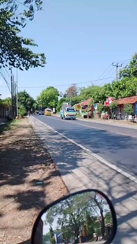 The video captures a sunny day on a paved road, likely in a rural or suburban area, as seen from the perspective of someone riding a motorcycle. The road curves slightly to the left, with a sidewalk or shoulder on the left side. Trees line the left side of the road, casting shadows across the path. On the right side of the road, there are buildings with red-tiled roofs, some appearing to be small shops or homes. Traffic includes several vehicles. A yellow bus is visible in the distance. A blue truck drives past. A black sedan with gold rims speeds by, followed by a red car. The mirror of the motorcycle reflects the trees and sky. The overall atmosphere is bright and clear.