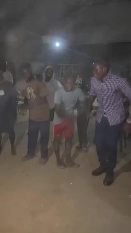 The video shows a group of five young people dancing indoors. The environment appears to be a dimly lit, unfinished space with a corrugated metal roof and rough concrete walls. Dust is visible on the floor. The person on the far right is wearing a purple and white checkered shirt and dark pants. Next to him, a person in a light blue t-shirt and red shorts is dancing. To their left, another person in a brown t-shirt and dark pants is dancing. Two other individuals are partially visible behind them, also dancing. The overall atmosphere suggests a casual gathering or celebration.