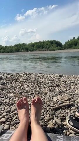 This is a first-person perspective video of a person relaxing by a river on a sunny day. The scene is set on a rocky shore with a calm river flowing in the background. Lush green trees line the opposite bank of the river. The sky is bright blue with scattered white clouds, suggesting a pleasant weather condition.

The primary subject is the person's bare feet, positioned in the foreground, resting on a patterned mat on the rocky shore. A pair of black and white sandals are visible to the right of the feet. The camera is focused on the feet and the immediate surroundings, looking out towards the river and the trees beyond.

The video captures a moment of relaxation and enjoyment of nature. The gentle movement of the water and the presence of natural elements evoke a sense of peace and tranquility. The lighting indicates it is daytime, likely midday or afternoon, given the bright sunlight.

While no specific activity is explicitly shown, the posture and setting suggest the person is lounging, sunbathing, or simply taking a break by the river. The overall mood is serene and leisurely. There is no visible text in the video. The location context provided is Bely Yar, Russia.