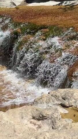 A close-up video captures a small waterfall cascading over rocks and moss. The water is clear and frothy, creating a dynamic spray. The surrounding rocks are light-colored and textured, with some patches of green moss and reddish-brown sediment visible on the upper rocks. The lighting suggests it is daytime, with bright sunlight illuminating the scene.