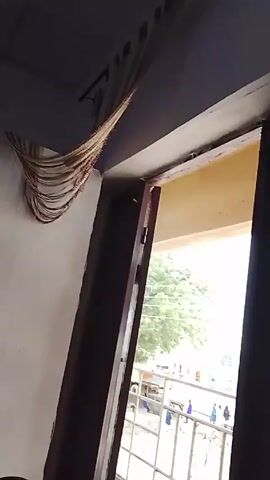 Here's a forensic description of the video:

The video shows a view from inside a room, looking out through a partly opened window. The window overlooks a street with a simple metal railing balcony. People are visible walking in the street, along with a vehicle. 

The camera then pans downwards to show a laptop sitting on a bed. The laptop screen displays what appears to be a photo editing or organization application.  A person is lying on the bed next to the laptop.
