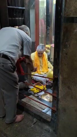 Two men are in a dimly lit, enclosed space, possibly a temple or shrine. In the foreground, facing away from the camera, a man in a light-colored shirt and brown pants stands with his left hand reaching out. He is holding a red cloth in his right hand. His left foot is bare. Behind a metal barrier, a man wearing a bright yellow garment and a matching scarf is seated on the floor. He has short gray hair and appears to be gesturing with his hands. In front of him are several yellow flower garlands and a metallic plate. The environment appears to be made of rough stone and metal. The lighting is low, casting shadows.