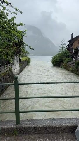 The video shows a turbulent, muddy river flowing through a village. The riverbanks are lined with stone walls and bordered by buildings and lush green trees. In the background, a large body of water, likely a lake, is visible, with misty mountains rising behind it. The sky is overcast and appears to be raining, contributing to the murky color of the water. A green metal railing is in the foreground, suggesting the footage is captured from a bridge or walkway overlooking the river. The overall atmosphere is somber and possibly indicative of heavy rainfall or flooding.
