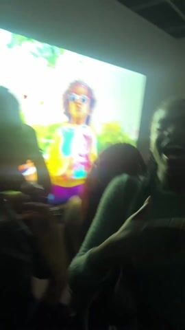 The video shows a group of people in a dark room, illuminated by a bright TV screen. The screen displays an animated scene with bubbles. A woman in the foreground is laughing and appears to be holding something in her hand, possibly a cigarette. Other individuals are partially visible in the background.