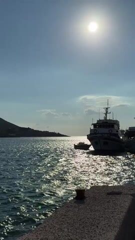 The video shows a body of water with a boat docked at a pier. The sun is shining brightly in the sky, creating a reflection on the water. The sky is clear with a few clouds. The water appears calm, with gentle waves. The boat is white and appears to be a pleasure craft or a small ferry. The pier is made of concrete and has a bollard for tying up boats. In the distance, there is a hillside covered with vegetation. The overall scene is peaceful and scenic, suggesting a summer day.