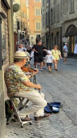 A street musician plays the violin while sitting on a folding chair on a cobblestone street. He is wearing a white hat, a floral shirt, and white pants. A violin case sits on the ground next to him. Several people are walking down the street in the background. The buildings on either side of the street are old and have shutters. The sun is shining, casting shadows on the ground.