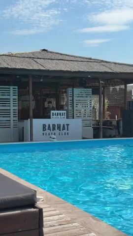 A wide shot shows a swimming pool in the foreground with a thatched-roof beach bar in the background. The beach bar has a white counter with "BARHAT BEACH CLUB" signage. There are several people visible behind the counter, appearing to be working. The water in the pool is a vibrant blue and reflects the sunlight. The sky is blue with some white clouds. The scene appears to be set during the daytime, likely in a warm climate given the beach bar and pool setting.
