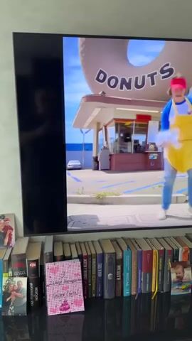 The video shows a television screen displaying two scenes. The first scene, from 00:00 to 00:02, features a person wearing a yellow apron over a blue shirt and jeans, dancing in front of a donut shop with a giant donut on top. The second scene, from 00:03 to 00:04, shows a person in a blue sweatshirt and red patterned pants dancing in a laundromat, with rows of washing machines in the background. Below the television, a shelf is filled with books, a few photographs, and a pink card with writing on it.