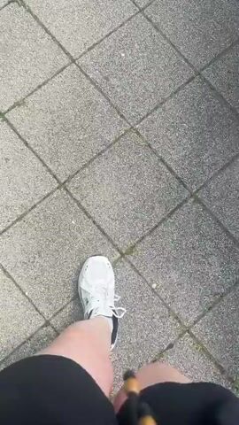 The video shows a first-person perspective of someone walking on a tiled sidewalk. The camera is positioned to focus on the person's right leg and foot, clad in a white athletic shoe with dark accents and black athletic shorts. The path is paved with square, light-colored tiles, with some sparse green vegetation pushing through the cracks. The lighting suggests it is daytime, likely sunny given the clear shadows and bright illumination. The scene appears to be outdoors, possibly in a park or a residential area given the urban but not overly busy environment suggested by the sidewalk. No text is visible in the video. The activity is simply walking, with no discernible emotion or specific event indicated.
