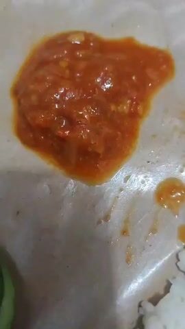 A close-up shot shows a plate of food served on brown paper. In the center of the frame is a dollop of red chili sauce. To the left of the sauce are two slices of cucumber and a piece of lettuce. To the right of the sauce is a piece of fried chicken. Below the sauce and to the right is a pile of white rice. In the background, a bottle of water is visible. The lighting is dim, and the focus is primarily on the food.