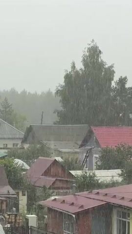 The video shows a view of a residential area during heavy rainfall. The sky is overcast and gray. Rain is visible as streaks falling from the sky, and it appears to be hitting the roofs of houses and the surrounding vegetation. Several houses with pitched roofs of various colors, including red and gray, are visible. Trees and bushes are present in the foreground and background, appearing wet from the rain. The overall atmosphere is of a downpour. No people or specific actions are clearly discernible.