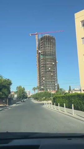 The video shows a tall, cylindrical skyscraper under construction, viewed from a moving car. A large crane is attached to the side of the building, extending upwards and to the right. The skyscraper is mostly exposed concrete and scaffolding, with some windows visible. The sky is clear blue. A road with cars parked on the side runs in front of the building. There are trees and bushes lining the road, and some power lines are visible overhead. The car appears to be driving on a sunny day, possibly in a residential or commercial area.