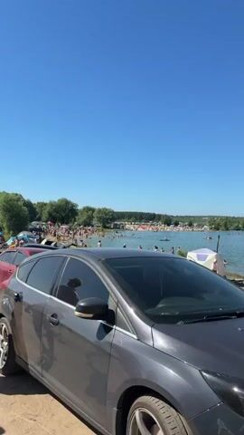 The video shows a crowded beach on a clear sunny day. Cars are parked along the sandy shore, with a red car in the foreground on the left and a gray car in the foreground on the right. A body of water, presumably a lake or sea, stretches out behind the cars, with numerous people swimming and relaxing on the beach and in the water. Tents and umbrellas are scattered across the beach, indicating a recreational gathering. The sky is a vibrant blue with no clouds visible. The overall scene depicts a bustling summer day at a popular beach destination.