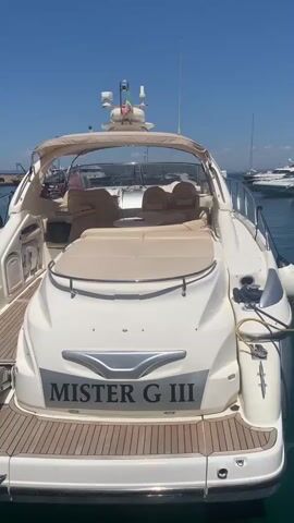 The video begins with a view of the stern of a white motor yacht named "MISTER G III". The yacht has a teak deck and a tan canopy over the cockpit area. Behind it, the video pans to the left, showing other boats docked in a harbor. There is a larger white and red boat with many fenders hanging off its side, and further in the background, a very large modern yacht with dark windows. The water in the harbor is clear and turquoise. The sky is a bright blue with no clouds. The scene appears to be a sunny day in a marina.