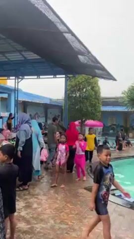 Here is a detailed description of the video:

The video shows a bustling outdoor swimming pool area. A mix of children and adults is present, some gathered near the pool's edge and others already in the water. The children wear colorful swimwear, and some adults are dressed in modest attire.

The pool itself is surrounded by tiled walkways and greenery, including palm trees and shrubs. There is a small bridge structure spanning a section of the pool, adding a decorative element. Overhead, a partial canopy provides shade to some of the area.

The weather appears overcast, suggesting it might be a cooler day or perhaps lightly raining. The overall atmosphere is lively and recreational.