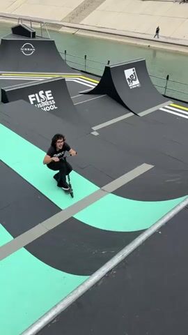A person wearing a black t-shirt, black jeans, and sneakers is riding a scooter on a large ramp course. They start by riding on a rail, then transition to a flat part of the course before going off-camera towards a ramp further ahead. The course is marked with black, gray, and mint green stripes. Logos for sponsors like FISE, Nissan, and Hurricane Parks are visible on various surfaces. The course is situated near a body of water with a walkway along the edge.