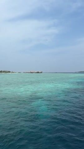 The video shows a serene view of the ocean under a blue sky with scattered clouds. The water transitions from a deep blue to a turquoise hue in the shallow areas, suggesting a coral reef beneath the surface. In the distance, there are small islands with buildings visible, possibly a resort. The camera appears to be on a boat, moving gently with the rhythm of the waves. There are no people visible in the shot.