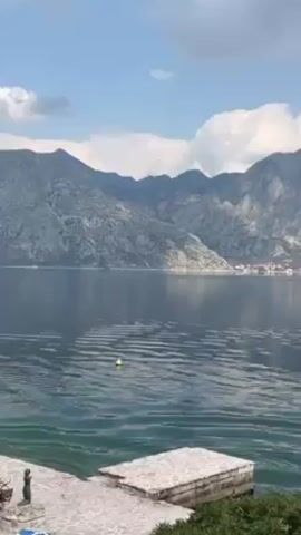 The video shows a view of a calm body of water, likely a lake or bay, surrounded by large, rocky mountains. The mountains are a mix of grey and white, possibly due to the presence of snow or light-colored rock formations. The water is a dark blue-green, with gentle ripples across the surface. A small yellow buoy bobs in the water. In the foreground, a stone dock or walkway is visible, partially obscured by greenery. The sky is light blue with scattered white clouds. The video pans slowly, focusing on the mountains across the water.