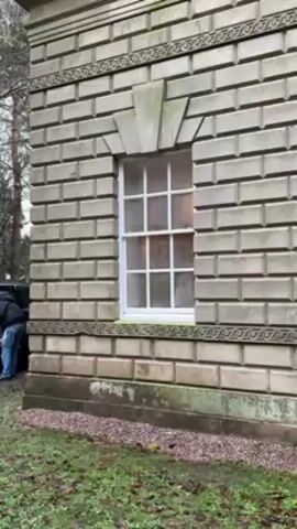 The video shows a person standing next to the open trunk of a dark grey SUV in a wooded area near an old stone building. The building is constructed of rectangular blocks and features a white-framed window on its side. A heart shape is drawn on the foggy glass of the window. The person wears a dark jacket, blue jeans, and a blue hat, and appears to be arranging items in the trunk. The background is filled with trees with bare branches, and the ground is covered with grass, moss, and some fallen leaves.