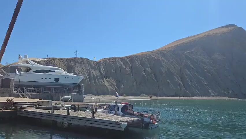 The video shows a harbor with several boats docked. In the foreground, a long wooden pier juts out into the water, with two boats moored alongside it. One of the boats is a small white boat with a red stripe, and the other is a larger vessel with an American flag painted on its side. In the background, a large white yacht is being lifted out of the water by a crane. The yacht is positioned on a platform above the water. Beyond the harbor, a large, barren mountain rises steeply from the coastline. The sky is clear and blue, and the water is a deep blue color. The overall scene is one of a busy harbor on a sunny day.