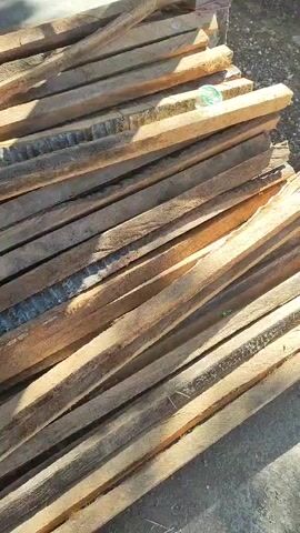 The video pans over a pile of wooden planks stacked horizontally. The planks appear to be rough and unfinished, with some showing signs of wear and tear. They are arranged in a disordered fashion, creating a layered effect. In the background, there is a glimpse of a concrete structure and some shadows cast by an unseen object. A green plastic bottle is visible on top of one of the planks. The lighting suggests an outdoor environment, possibly a construction site or a storage area. The camera movement is steady, creating a continuous visual flow across the wooden material.