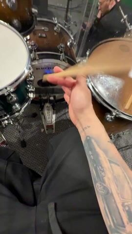 A first-person view video shows a person playing a drum set. The drummer is wearing black pants, and their left arm has a tattoo of a car. The drum set is a variety of brown, silver and teal colored drums. A drum pedal and cymbals are visible. In the background, another person wearing glasses can be seen sitting in a chair. The scene appears to be a music studio or practice room. The drummer throws a drumstick in the air and catches it.