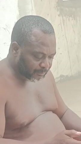 Here's a description of the video:
A medium shot shows a middle-aged, dark-skinned man with a graying beard sitting shirtless. He's looking down, seemingly focused on something in his hands. He has a receding hairline and a light dusting of gray in his beard. The background is a white or light-colored textured wall. The lighting is relatively even.