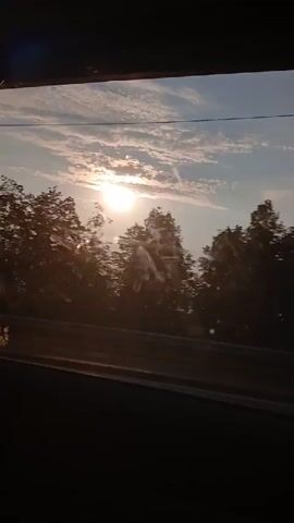 The video shows a view from a moving vehicle, likely a car. The camera is pointed out a window. In the foreground, the dark interior of the vehicle is visible, including the window frame and a thin, vertical object that may be a windshield wiper or a thin pillar. The middle ground features a road or highway, with visible lanes. Beyond the road, a silhouette of trees and hills is present. The sky is bright with the sun visible and shining through scattered clouds. Lens flares and dust particles are visible on the camera lens. The overall lighting suggests either sunrise or sunset.