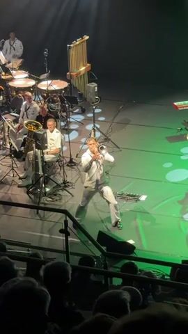 A trombonist in a white uniform plays a solo in the center of a stage. He is surrounded by other musicians playing brass and percussion instruments. The stage is lit with green and white lights, and there is an audience in the foreground.