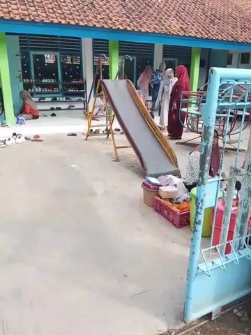The video shows a playground area outside a building. The building has a red tile roof, light blue trim, and green pillars. There is a shoe rack filled with shoes outside the building. Several people are visible, most wearing hijabs. A woman in a light-colored hijab and a young child in a dark outfit are standing near a slide. Another woman in a maroon hijab is standing nearby. There is a small slide in the center of the playground. Next to the slide, there are containers with goods. A woman wearing a hijab sits behind the goods. A blue metal gate partially obscures the view of the scene.  A woman sits near the shoe rack outside the building.