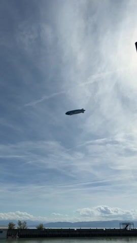 A blimp with the logo "ZF" and the text "Next Generation Mobility. NOW." written on its side flies through the sky. The blimp is white with blue accents and has a gondola underneath. The sky is blue with some scattered clouds. In the foreground, a body of water with a dock is visible.