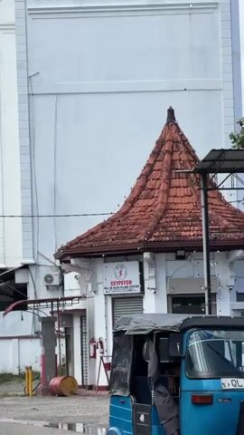 The video captures an exterior view of buildings in Matara, Sri Lanka, under an overcast sky. The primary subject is a large, white, multi-story building featuring the green-lettered sign "SUPIRI WINE STORES" on its upper section. In front of and slightly below this larger structure is a smaller, white building with a distinctive red-tiled conical roof. A sign on this smaller building reads "CEYPETCO M.A. DE SILVA FILLING STATION." Visible nearby are a red barrel, two fire extinguishers, and some metal scaffolding or posts. The camera slowly pans upwards, first focusing on the "SUPIRI WINE STORES" sign and then continuing to show only the cloudy sky, filled with white and grey clouds. No people or specific activities are shown; the video presents a static architectural scene on a cloudy day.