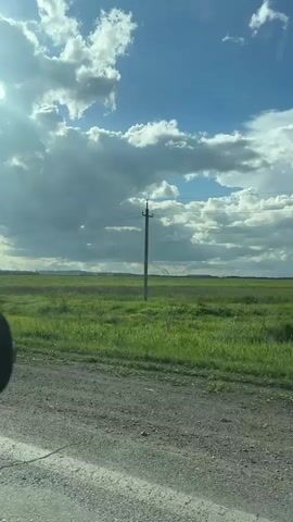 A landscape view from a moving vehicle shows a vast green field under a partly cloudy sky. In the distance, several industrial structures with what appear to be smoke or steam plumes rise above the horizon. A metal guardrail runs along the bottom of the frame, separating the field from a dirt road in the foreground. A utility pole with power lines is visible on the left side, passing by as the vehicle moves. The sun is shining, casting shadows and highlighting the clouds. The overall impression is of an open, rural landscape with industrial activity in the far background.