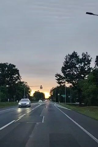 The video shows a view from a car driving down a road lined with trees. The sky is overcast, with the sun visible low on the horizon, suggesting it is either sunrise or sunset. There are other cars on the road, and pedestrian crossing signs are visible. The road has lane markings, and there are streetlights along the sides. The overall impression is of a journey through a semi-rural or suburban area at dusk or dawn.