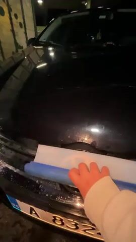 Here's a description of the video:

The video shows a person cleaning the front grill of a black Volkswagen car. The car is parked in what appears to be a car wash area with an awning and overhead lighting. The person uses a blue squeegee to wipe down the front of the car, starting with the VW logo on the grill.  The car's license plate, with Bulgarian markings, is visible (A 8324 HP). The surroundings are dark, suggesting it's nighttime.