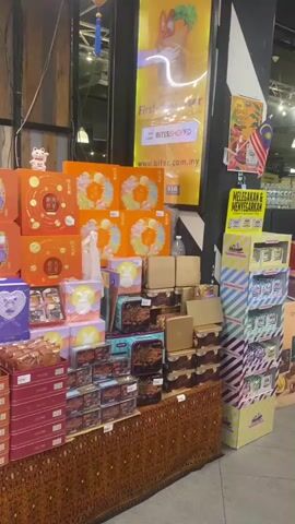 The video shows a display of gift boxes, likely for a holiday like the Mid-Autumn Festival, in a retail setting. The boxes are stacked high on tables and shelves, with a variety of colors and designs. Some boxes are orange, red, or gold, while others are purple or pink. Many of the boxes feature intricate patterns and illustrations, including floral designs, animals, and traditional Chinese motifs. Some boxes are open, revealing smaller packages or individual items inside, which appear to be pastries or mooncakes. The background shows a store interior with lighting, decorations like tinsel and paper lanterns, and other merchandise on shelves. There are no people visible in the video, and no actions or interactions are taking place beyond the static display of goods. The overall impression is a festive and abundant presentation of holiday gifts.