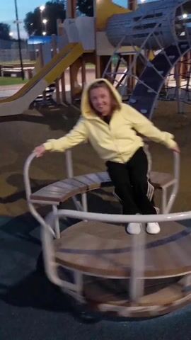 A young woman wearing a yellow hoodie and black pants is sitting on a merry-go-round in a playground. She is smiling and looking at the camera. The merry-go-round is spinning. In the background, there is a playground structure with slides and climbing equipment. The sky is dark, indicating it is either dusk or night.