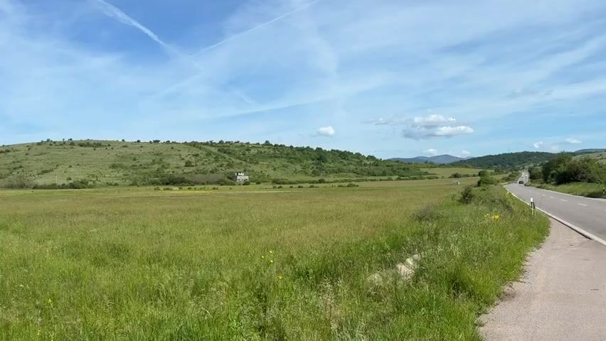 The video shows a wide shot of a vast, green, grassy field under a blue sky with scattered clouds and contrails. A paved road runs along the right edge of the frame, curving into the distance. In the background, a gently sloping hill covered in green vegetation and sparse trees rises. A small, light-colored building, possibly a house or shed, is visible on the hillside. The overall impression is a rural, natural landscape on a clear day. There are no people or animals visible in the frame. The camera appears to be stationary.