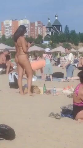 The media file depicts a daytime scene at a sandy beach, likely during warm weather. The sun is shining, indicated by the bright lighting and shadows. In the foreground, a woman wearing a minimal bikini stands facing away from the camera, holding a mobile phone. Other individuals are visible in the background, some sunbathing on lounge chairs or towels, while others are standing or interacting. A young child in a light blue outfit and a white cap is also present.

Behind the beachgoers, several beach umbrellas provide shade. Further in the background, there is a cluster of apartment buildings and a distinctive church with green domes. The overall atmosphere appears relaxed and casual, typical of a beach outing. There are no discernible signs or text visible in the footage. The scene suggests a popular public beach in a populated area.