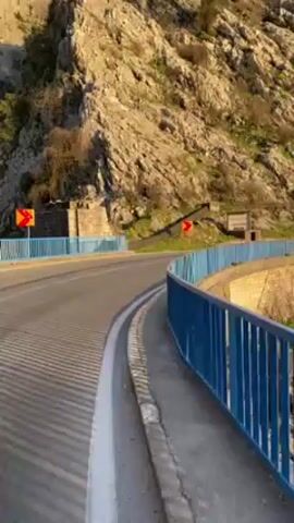 The video shows a blue-railed bridge over a rocky shore leading to a body of water, with mountains visible in the background. The camera pans from the road and bridge to reveal a riverbed and a lake, capturing the early morning sun reflecting off the water. The scene includes natural elements like rocks, trees, and mountains, under a clear sky.