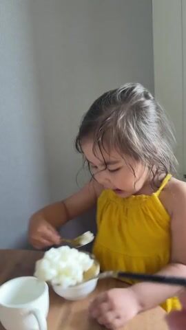 A young child with wet dark brown hair and a yellow sundress is sitting at a wooden table. The child is holding a small golden spoon in their right hand. A hand from outside the frame is holding a small black and gold spoon filled with white food towards the child's mouth. The child bites the food off the spoon. The child has food on their mouth and chin. A small white bowl is on the table in front of the child, and a small white cup is to the left of the bowl. The background is a plain light gray wall.