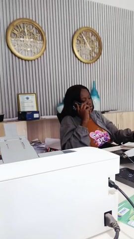 The video begins in what appears to be an office. A woman is seated at a desk, talking on her smartphone and looking at a computer monitor. The desk is cluttered with a printer, papers, and office supplies. There are two gold decorative wall clocks behind her. The camera then pans to the right, showing the back of the computer monitor. The view expands to reveal a waiting area with light blue sofas, a silver and marble coffee table, and a decorative patterned wall. There is a staircase visible in the background. The room has white tiled floors with a black and white patterned border. The video appears to be a quick tour of the office and waiting area.