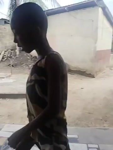 A young, dark-skinned child with short dark hair, wearing a yellow, black, and white patterned sleeveless dress, walks across a tiled porch. The porch features irregular, multi-colored tiles in shades of brown, white, and black. The child walks from the right side of the frame towards a light pink wall, where a blue-framed window and a dark wooden paneled door are located. The child approaches the window and stands facing it, looking out. The background shows a dirt yard with scattered debris, including tires and bricks, a palm tree, and other simple buildings under an overcast sky.